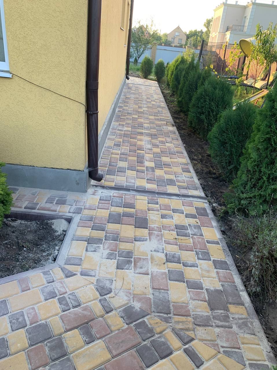 Ukończony chodnik z kostki brukowej w odcieniach żółci, brązu i szarości, biegnący wzdłuż ściany domu z brązową rynną i obsadzony tujami.