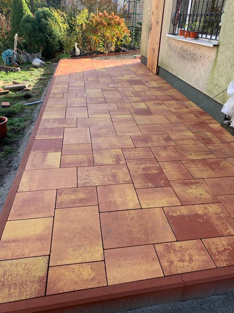 Ułożona, czerwono-żółta kostka brukowa o nieregularnym kształcie, tworząca ścieżkę przy budynku z drewnianymi drzwiami i oknem z doniczkami, widok z góry.