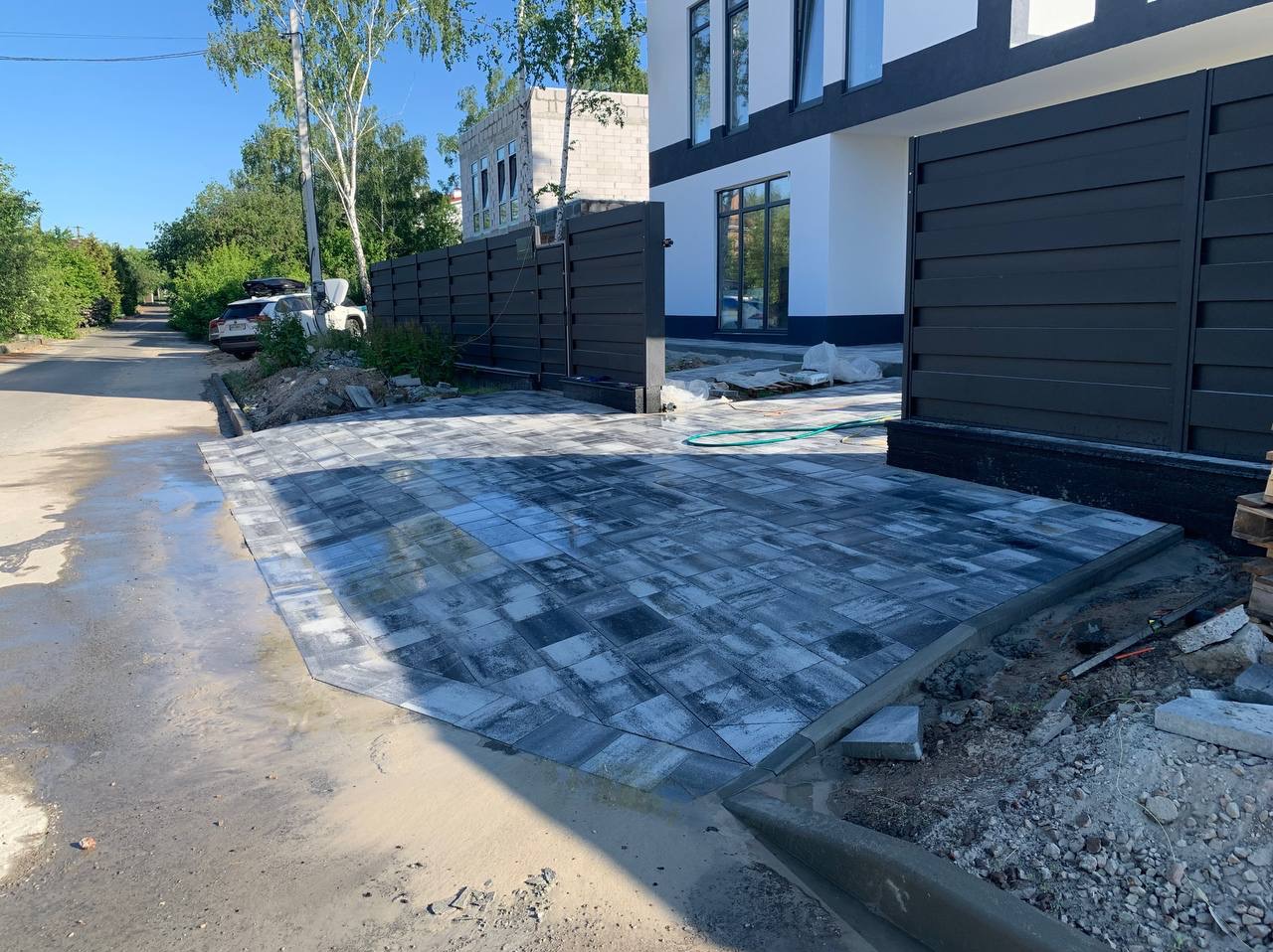 Ułożona kostka brukowa w odcieniach szarości przed nowoczesnym domem z czarnym ogrodzeniem; widoczne mokre plamy i pozostałości piasku.