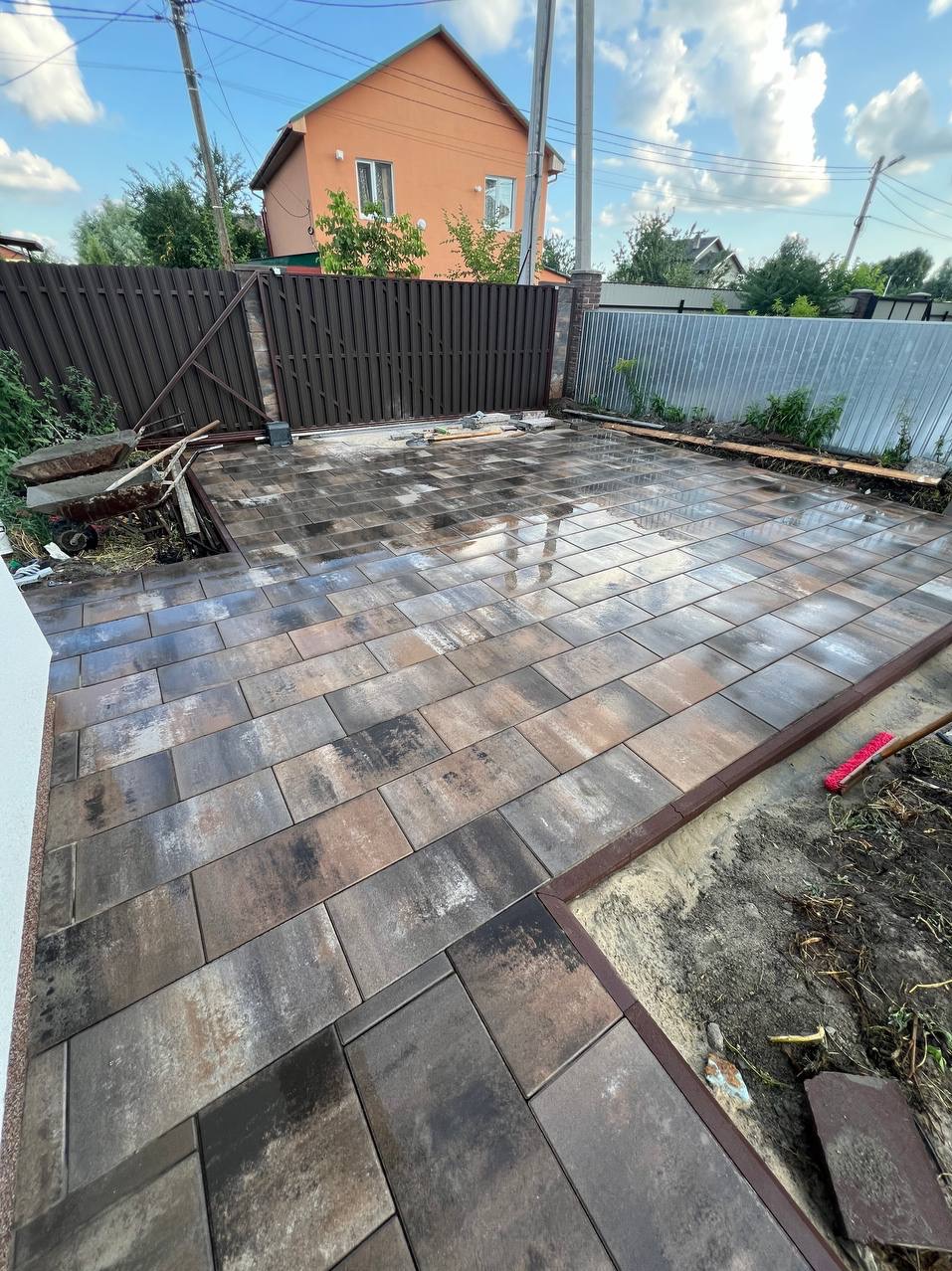 Ułożona, mokra kostka brukowa w odcieniach brązu i szarości na podwórku, z widocznym obramowaniem i fragmentem ogrodu w tle, z brązową bramą wjazdową i metalowym ogrodzeniem.