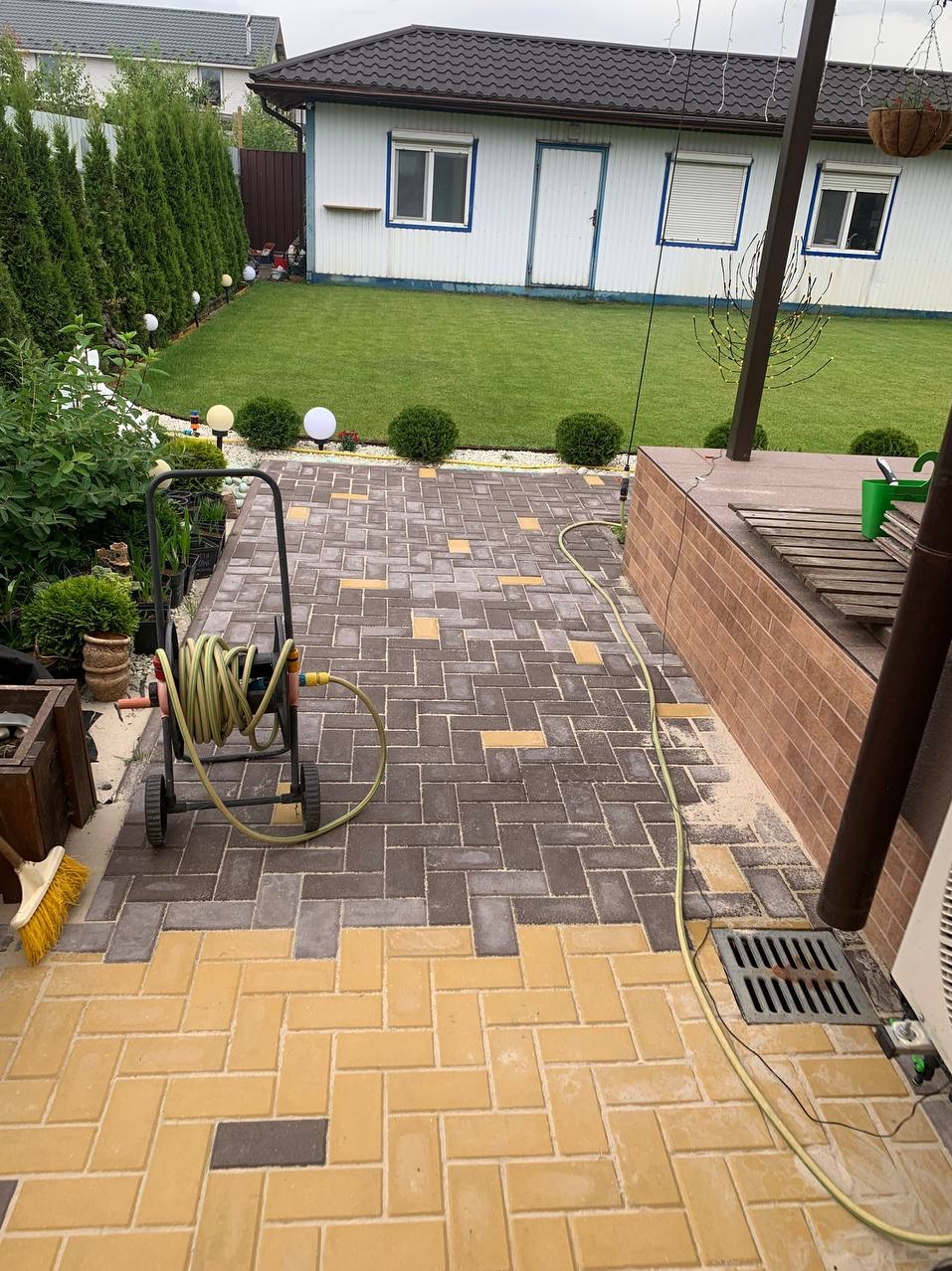 Ułożona kostka brukowa w dwóch kolorach, żółtym i szarym, z widocznym wózkiem na wąż ogrodowy i odpływem liniowym, na tle zadbanego ogrodu z trawnikiem i budynkiem gospodarczym.