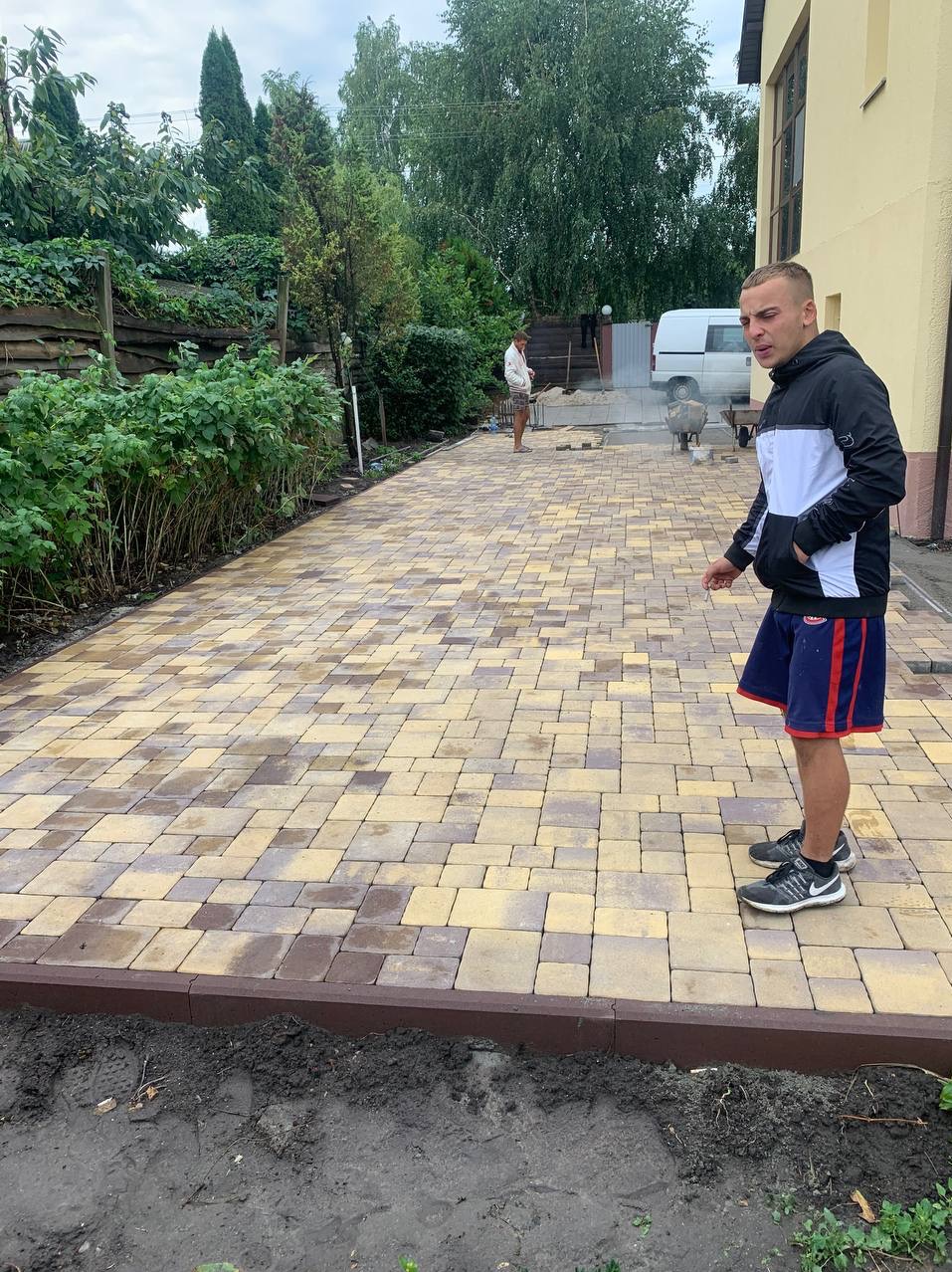 Ułożona kostka brukowa w odcieniach beżu i brązu, obrzeża z ciemnobrązowej kostki, osoba w sportowym stroju przygląda się pracy, w tle zaparkowany biały samochód dostawczy oraz druga osoba przy pracy.