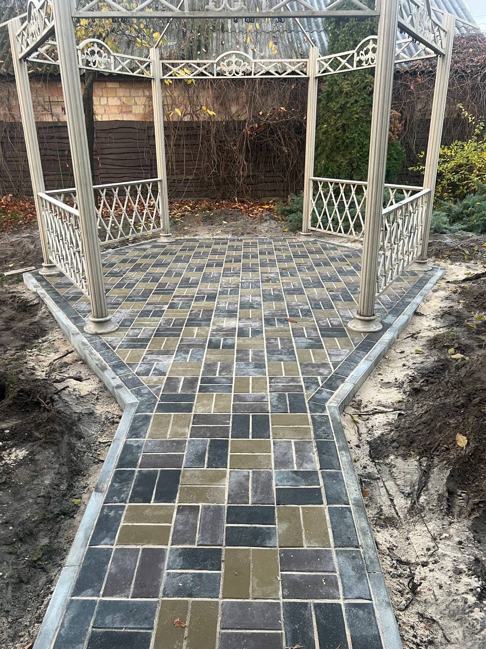 Nowo ułożona, wielobarwna kostka brukowa w kształcie prostokątów, tworząca ścieżkę i plac pod metalową altaną ogrodową z dekoracyjnymi balustradami, widoczne ślady prac ziemnych.