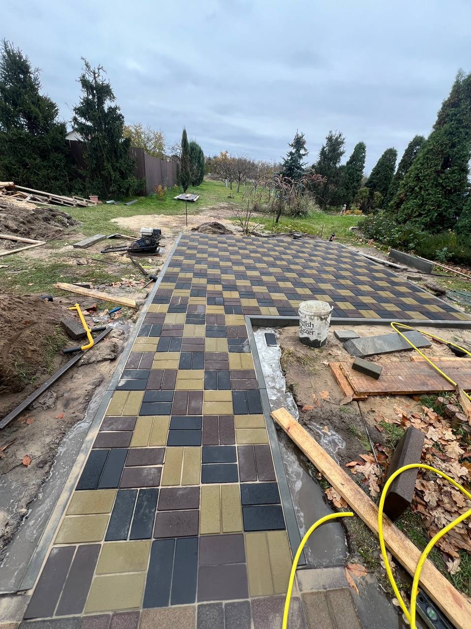 Ułożona kostka brukowa w szachownicę z brązowych, czarnych i beżowych elementów, widoczny fragment obrzeża i narzędzia, tło z drzewami i pochmurnym niebem.