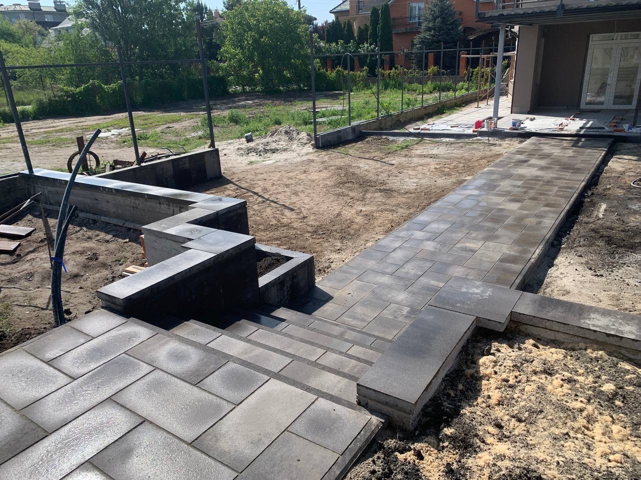 Układanie ciemnoszarej kostki brukowej na ścieżce i schodach w ogrodzie w Krosno Odrzańskie, widoczne elementy murku z kostki i przygotowany teren.