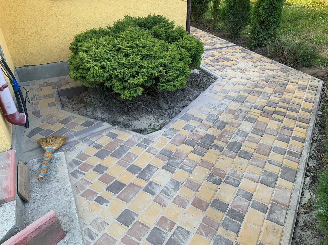 Ścieżka z kostki brukowej w odcieniach szarości, żółci i brązu, obok budynku z zielonym krzewem i miotłą opartą o ścianę.