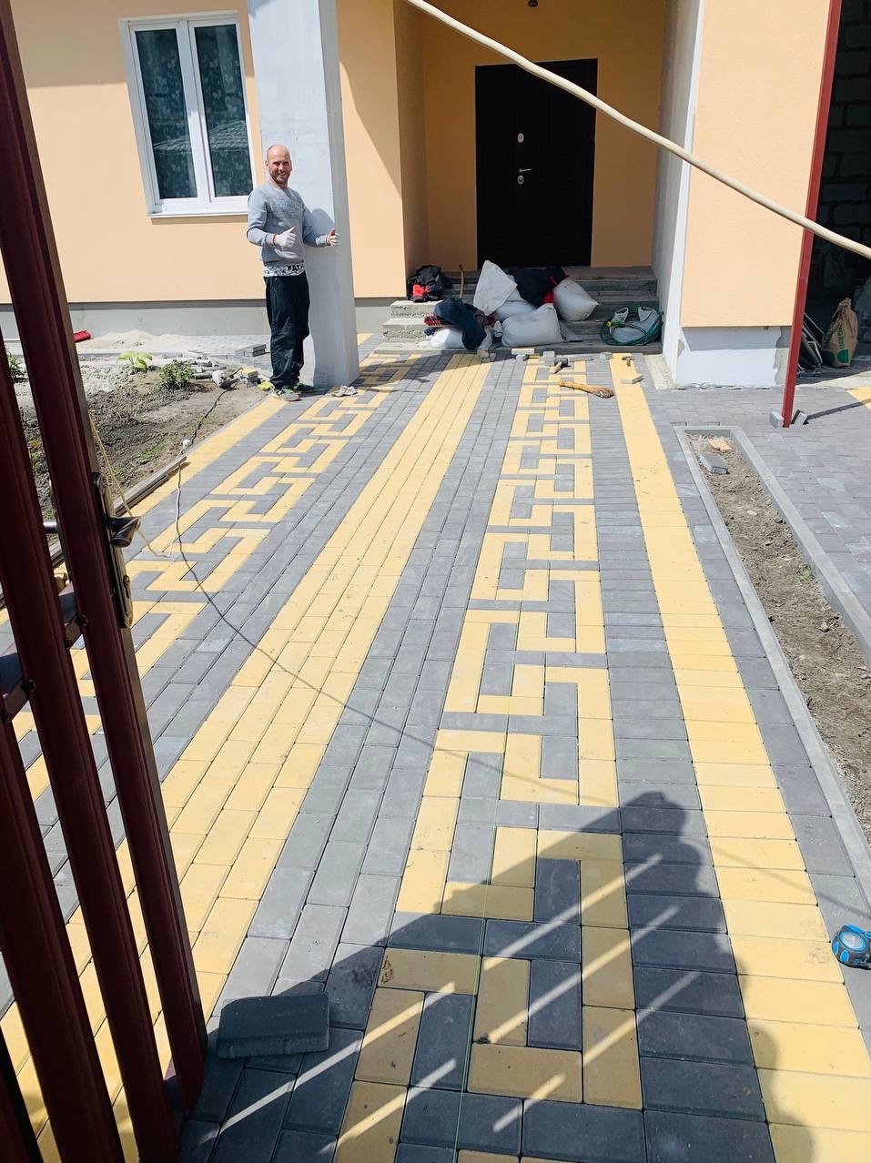 Ułożona kostka brukowa w geometryczny wzór z żółtych i szarych elementów przed wejściem do domu; pracownik budowlany pozuje z uniesionymi kciukami, obok leżą worki z materiałami budowlanymi.