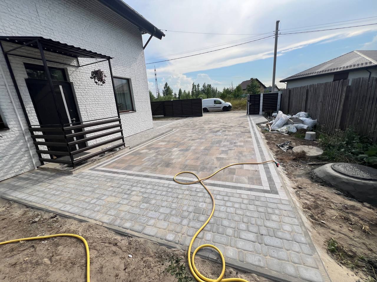 Ułożona kostka brukowa na podjeździe domu jednorodzinnego, z widocznym fragmentem elewacji z białej cegły, żółtym wężem ogrodowym i zaparkowanym białym vanem w tle.