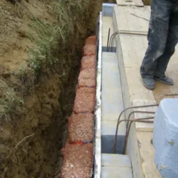 Wykonanie drenażu w wykopie, zabezpieczone workami z kamieniami, widoczne zbrojenie i elementy betonowe fundamentu, pracownik w ubrudzonych spodniach w tle.