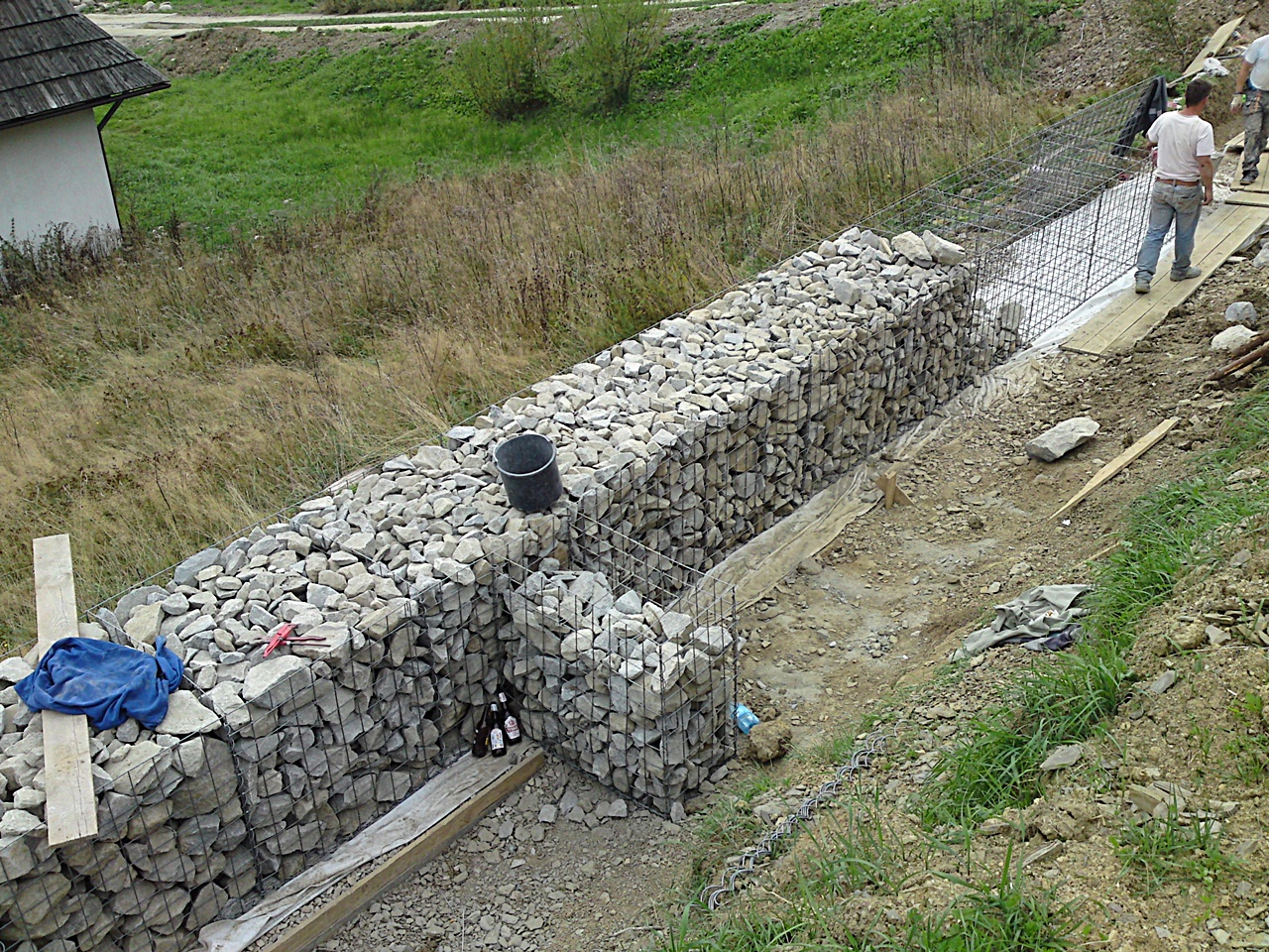 Budowa muru oporowego z gabionów wypełnionych kamieniem, widoczne elementy konstrukcji z siatki drucianej, wiadro i butelki na kamieniach, teren w trakcie prac ziemnych.