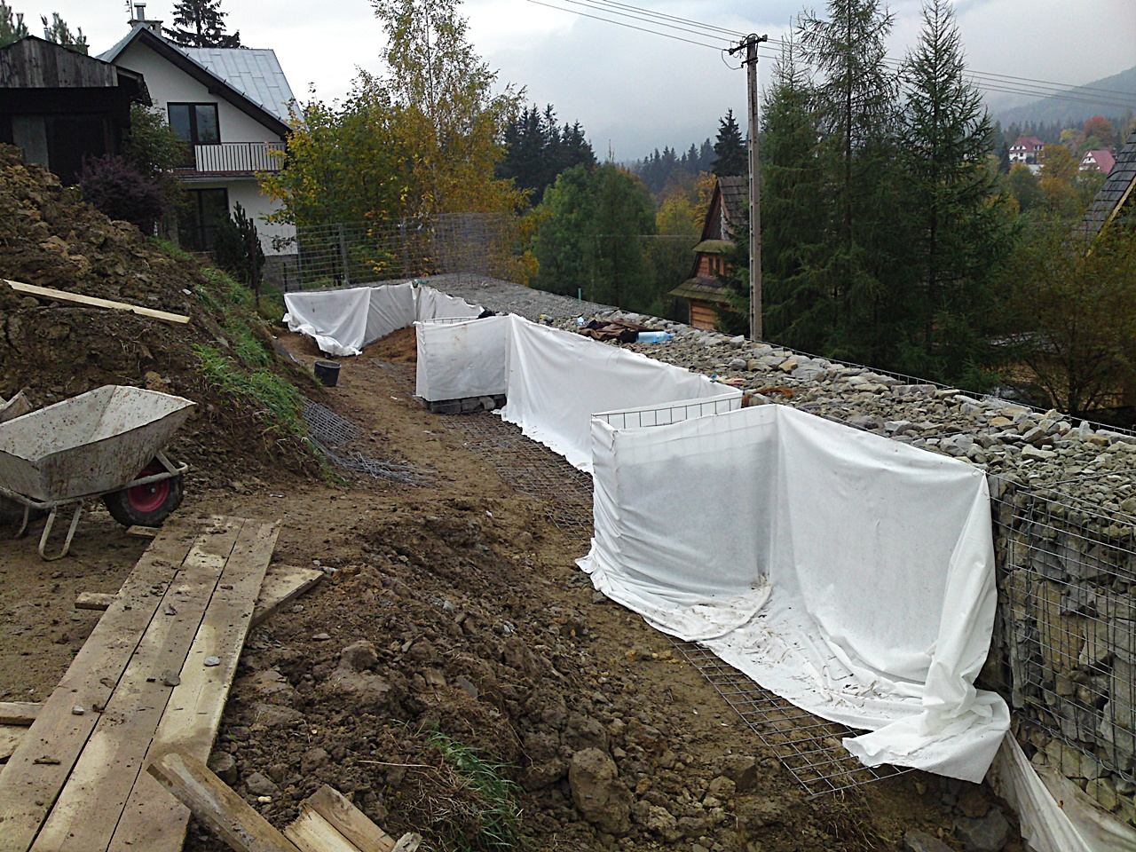 Budowa gabionów na zboczu, widoczne kosze z siatki wypełnione kamieniami, częściowo zakryte białą geowłókniną, taczka i deski na pierwszym planie, w tle domy i drzewa