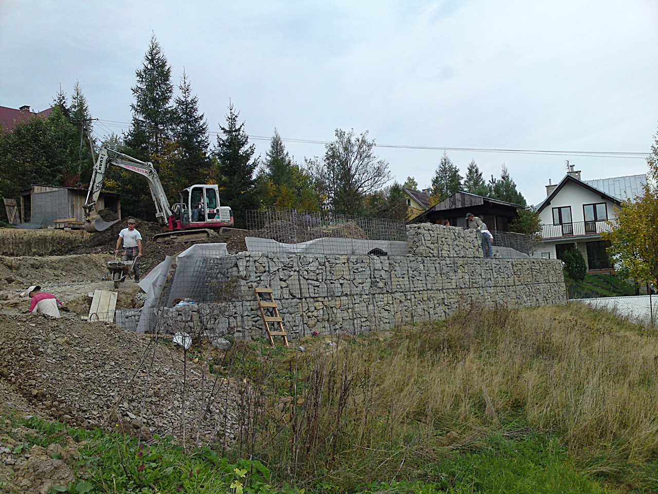 Budowa muru oporowego z gabionów na nierównym terenie, widoczny pracujący sprzęt budowlany, w tle domy jednorodzinne.