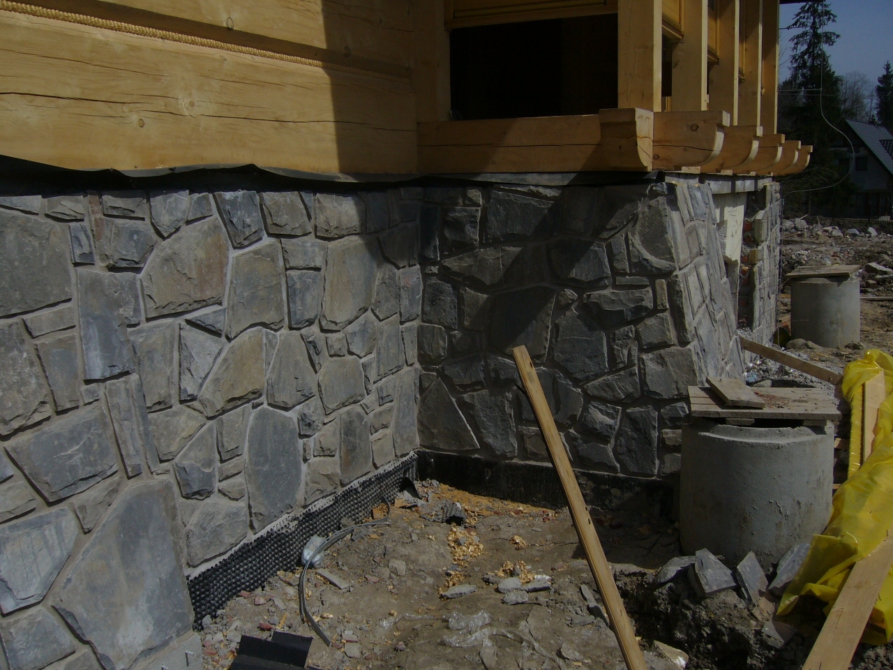 Fragment ściany budynku w trakcie budowy, wykończony kamieniem elewacyjnym. Widoczna hydroizolacja przy fundamencie oraz elementy konstrukcji drewnianej. Na pierwszym planie gruz i materiały...