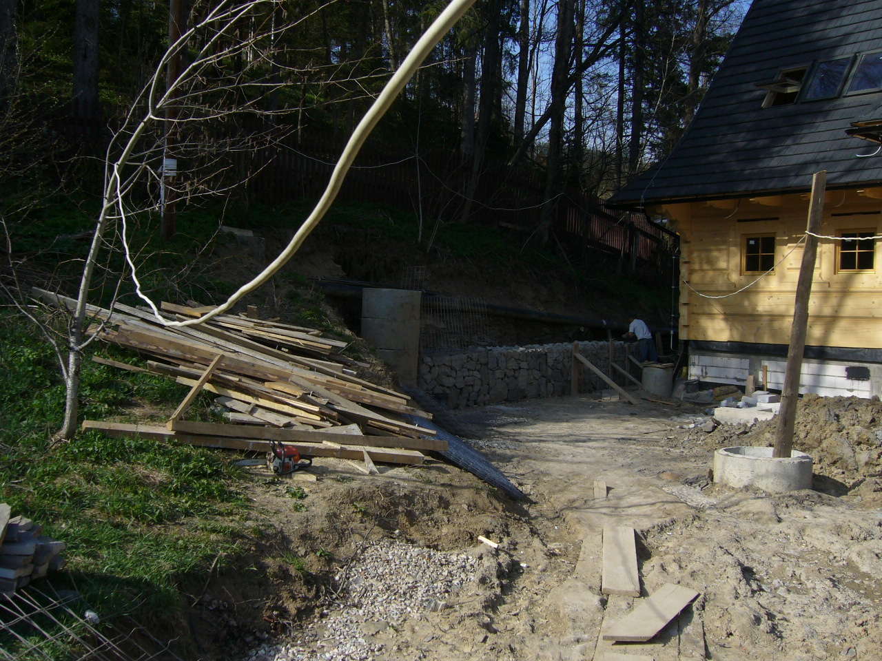 Budowa domu z bali: widoczne fundamenty, częściowo wykonane umocnienie skarpy z kamienia, rury drenażowe, stos drewna i piła łańcuchowa na zboczu.