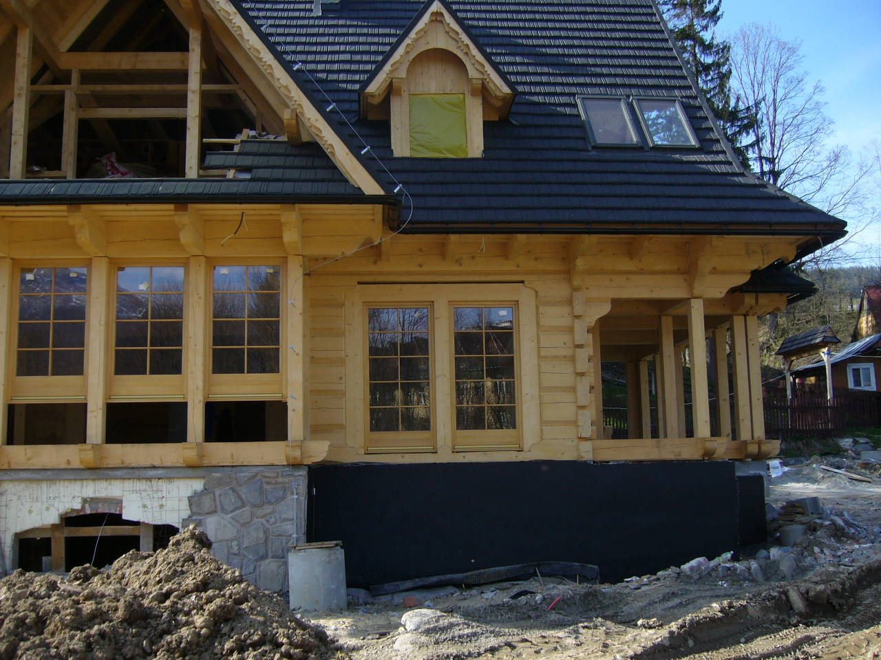Surowy stan drewnianego domu z czarną dachówką, widoczne okna z szybami, niezabezpieczone otwory w fundamentach oraz sterta ziemi przed budynkiem.