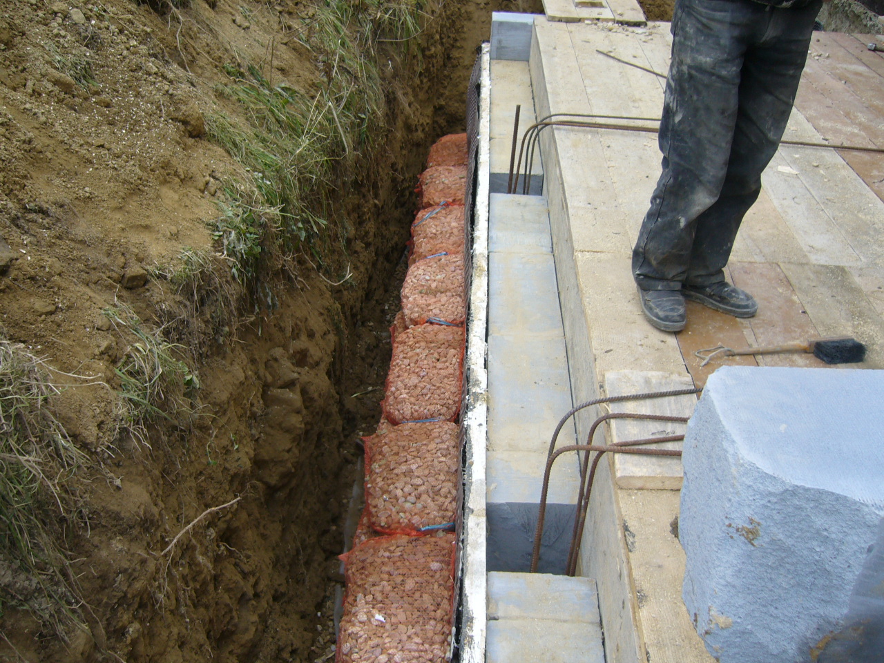 Wykonanie drenażu w wykopie, zabezpieczone workami z kamieniami, widoczne zbrojenie i elementy betonowe fundamentu, pracownik w ubrudzonych spodniach w tle.