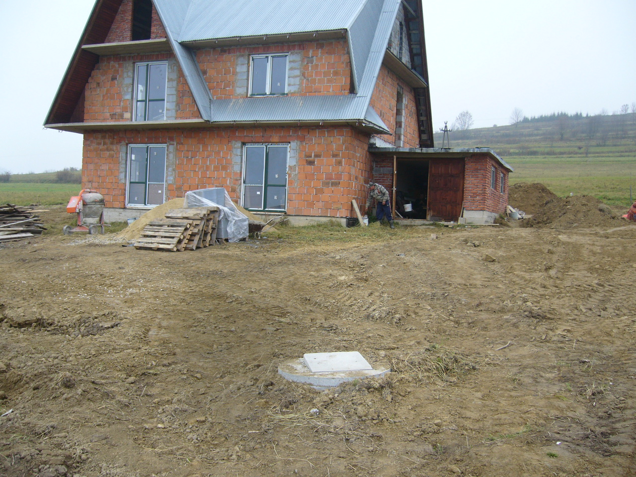 Budowa domu murowanego z czerwonej cegły, widoczne okna z zielonymi ramami, sterta piasku, palety, betoniarka, ziemia wokół budynku i wzgórze w tle.