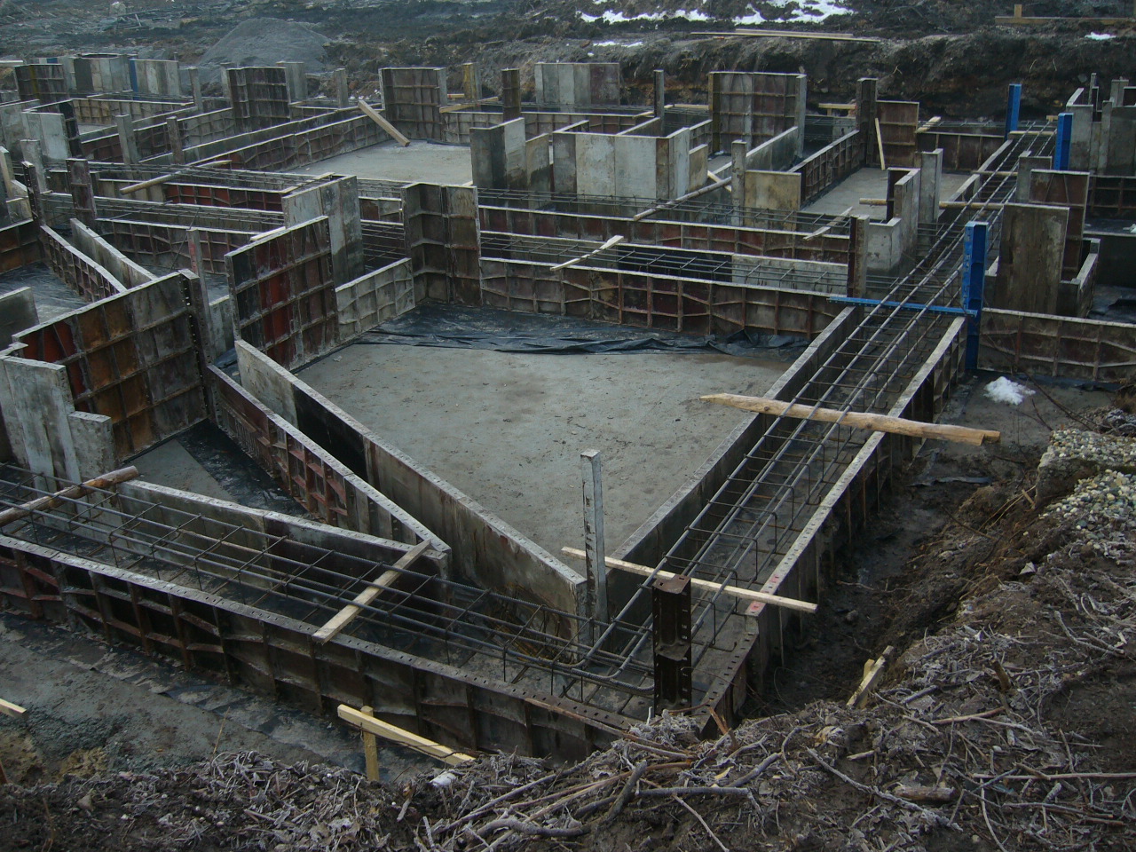 Zalane betonem szalunki fundamentowe w ziemi, widoczne zbrojenia i deski podtrzymujące konstrukcję. Częściowo zasypane ziemią.