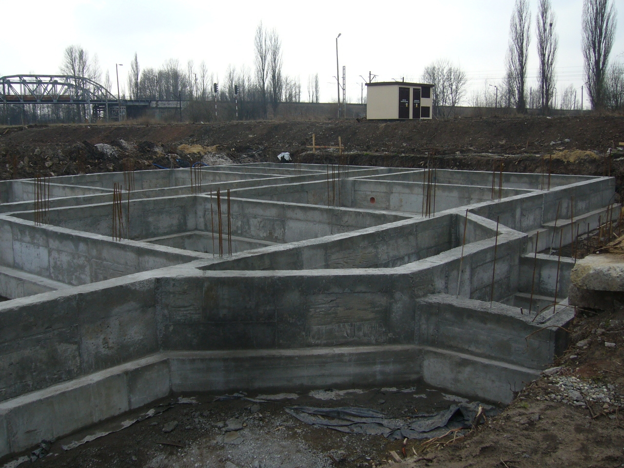 Zalane betonem fundamenty domu z wystającymi prętami zbrojeniowymi, widok z poziomu gruntu, w tle most kolejowy i budynek stacji transformatorowej.