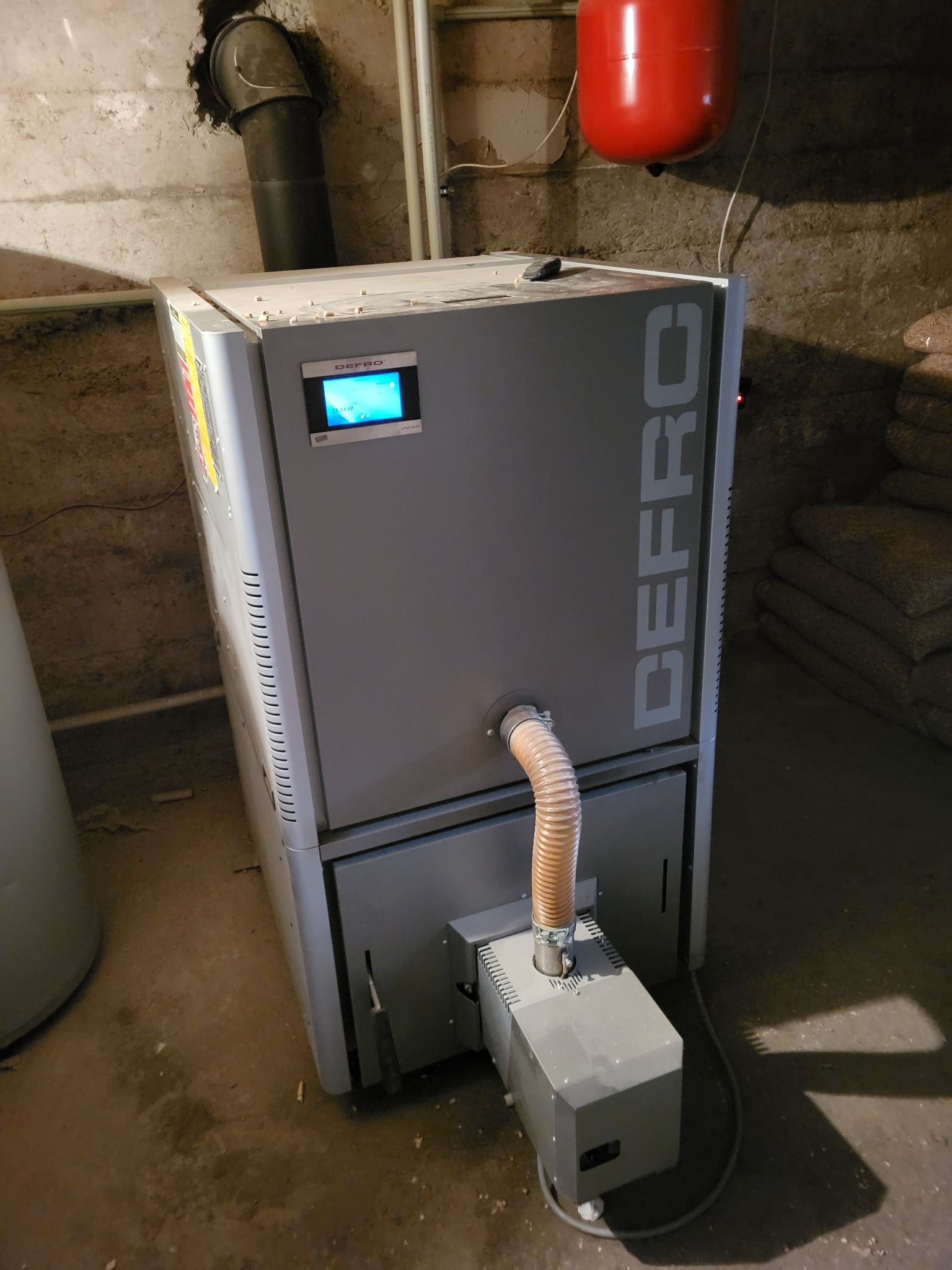 Szary piec Defro z podajnikiem i rurą spalinową w kotłowni. Widoczny zbiornik wyrównawczy i worki z pelletem w tle. Nierówne ściany i podłoga.