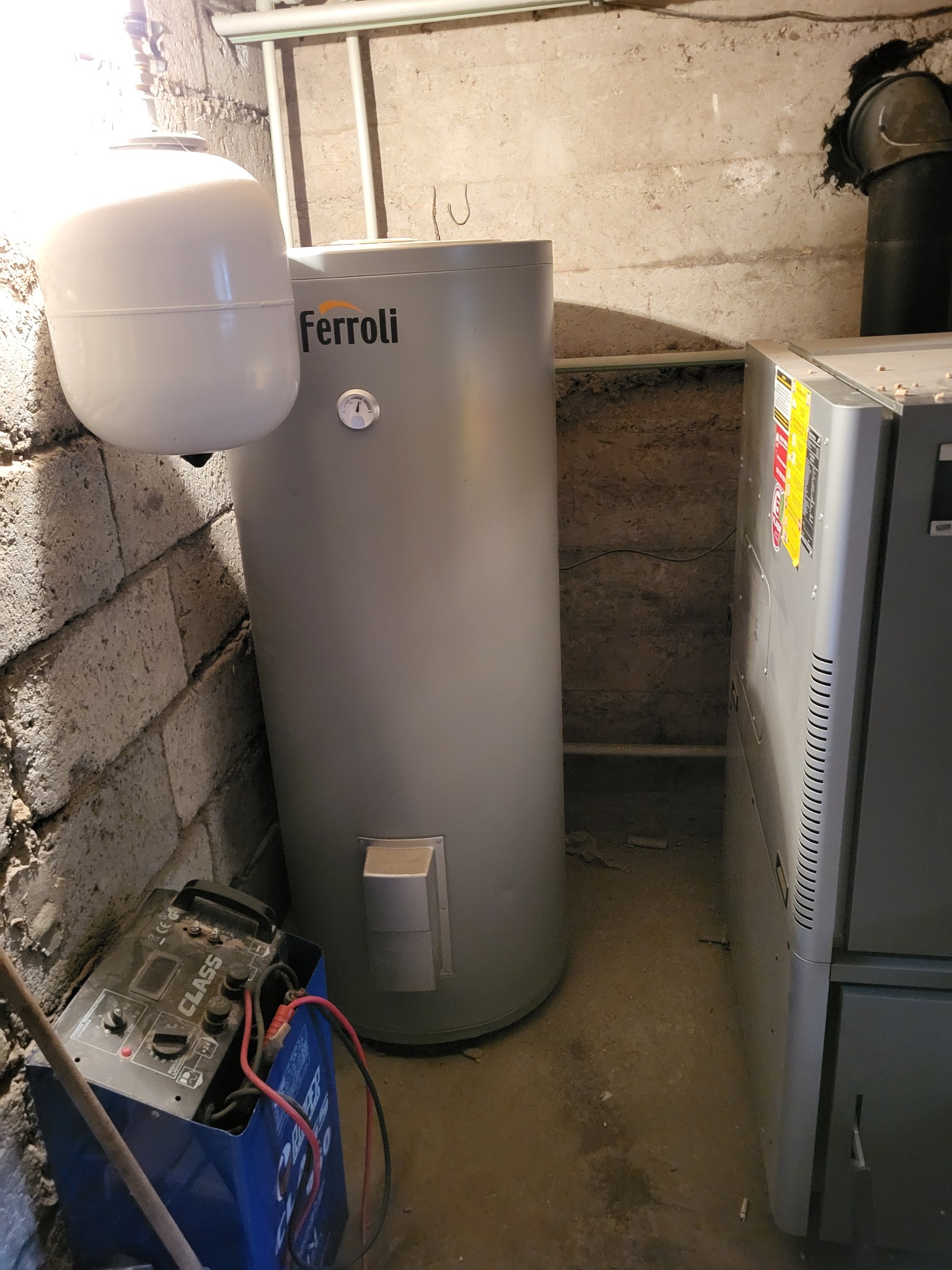 Szary, cylindryczny bojler Ferroli z białym zbiornikiem wyrównawczym, stojący w piwnicy przy betonowej ścianie i ładowarce akumulatorów.