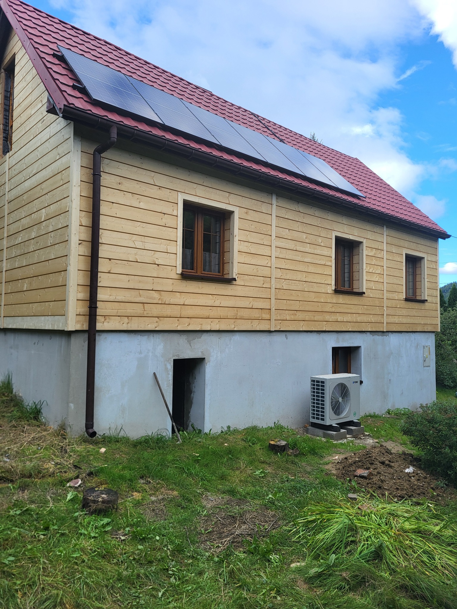 Dom z drewnianą elewacją i panelami słonecznymi na czerwonym dachu, z klimatyzatorem na zewnątrz. Widoczne okna i fragment ogrodu.