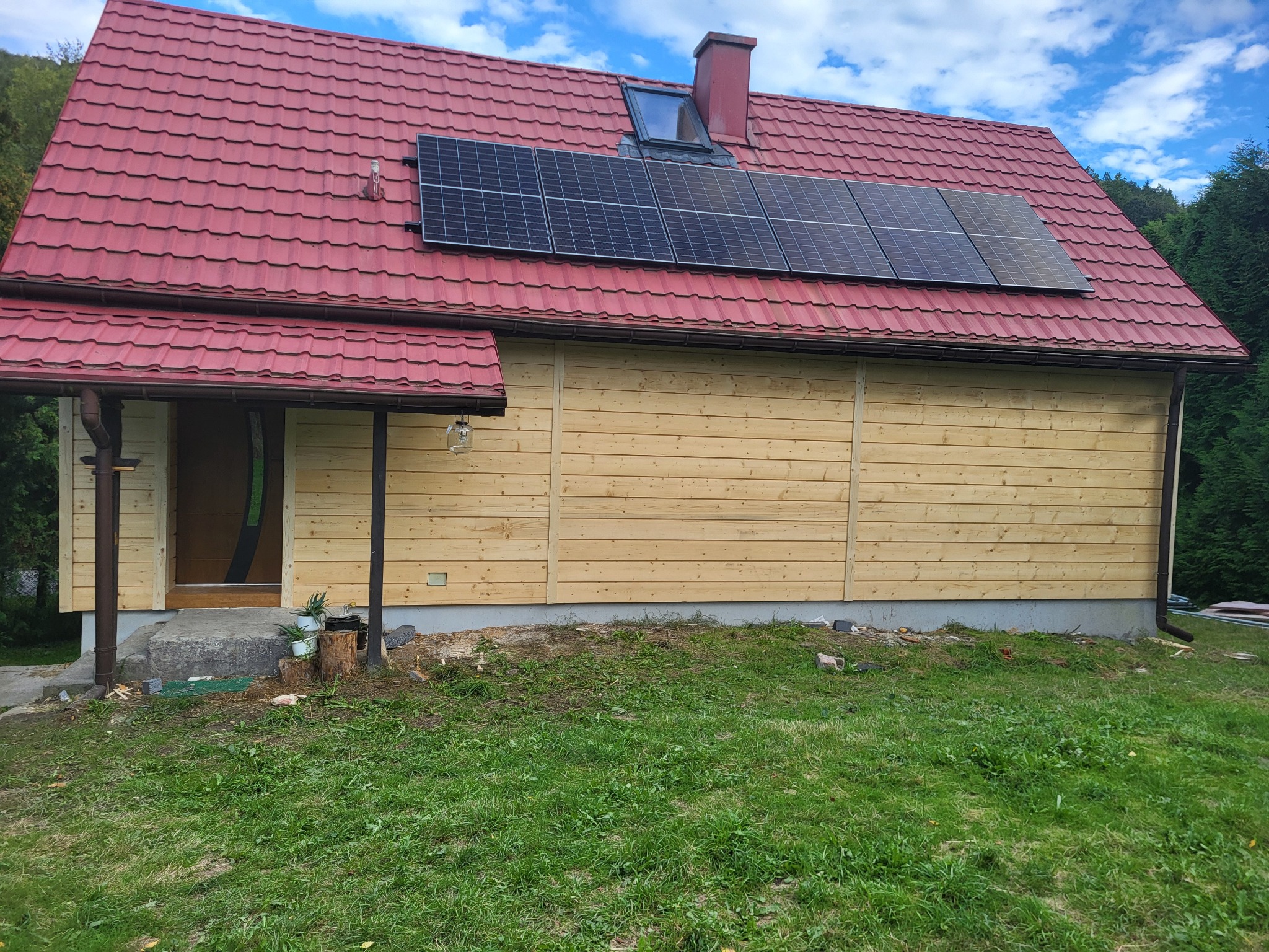 Dom z czerwoną dachówką i panelami słonecznymi. Ściany wyłożone jasnym drewnem. Widoczny fragment trawnika i drzew. Ujęcie z poziomu gruntu.