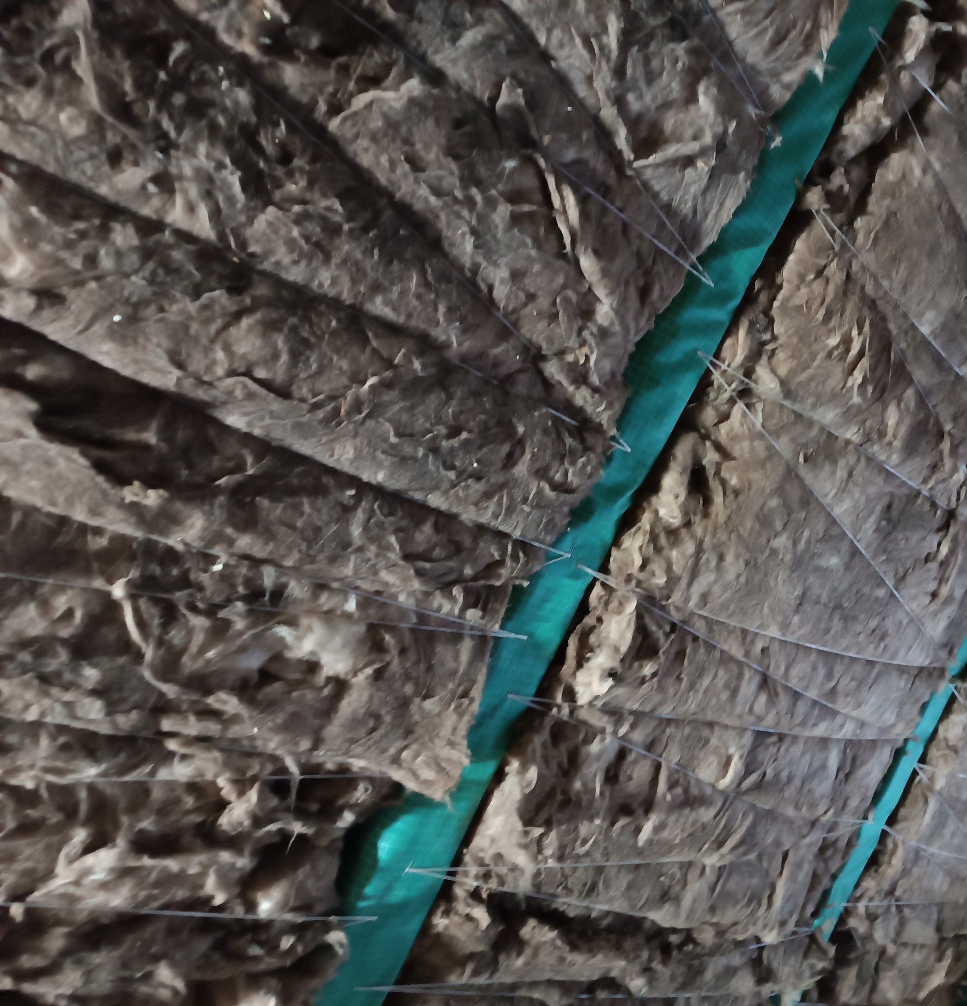 Warstwy izolacji z wełny mineralnej, zabezpieczone siatką i zieloną folią, tworzące strukturę ocieplenia. Widoczne detale mocowania i faktura materiałów.