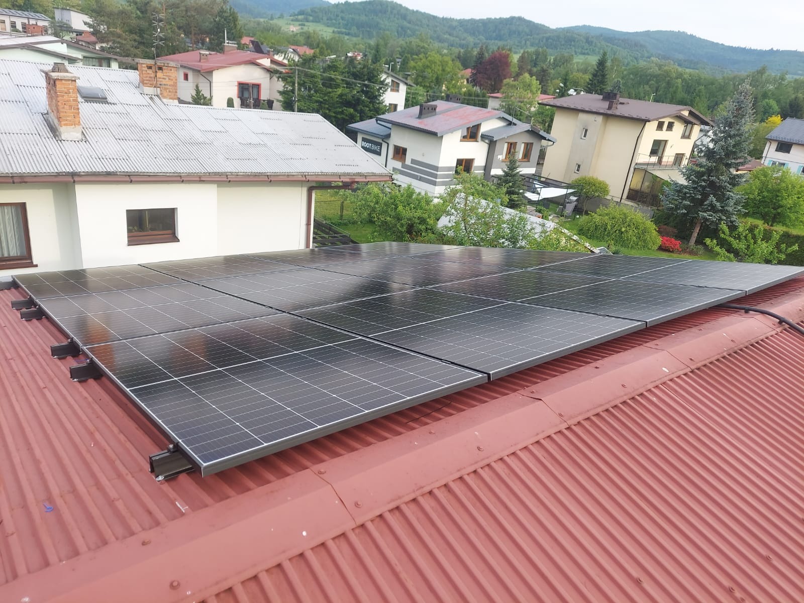 Panele słoneczne na czerwonym, blaszanym dachu domu jednorodzinnego, w tle zabudowa mieszkalna i zalesione wzgórza. Widok z góry, lekki kąt.