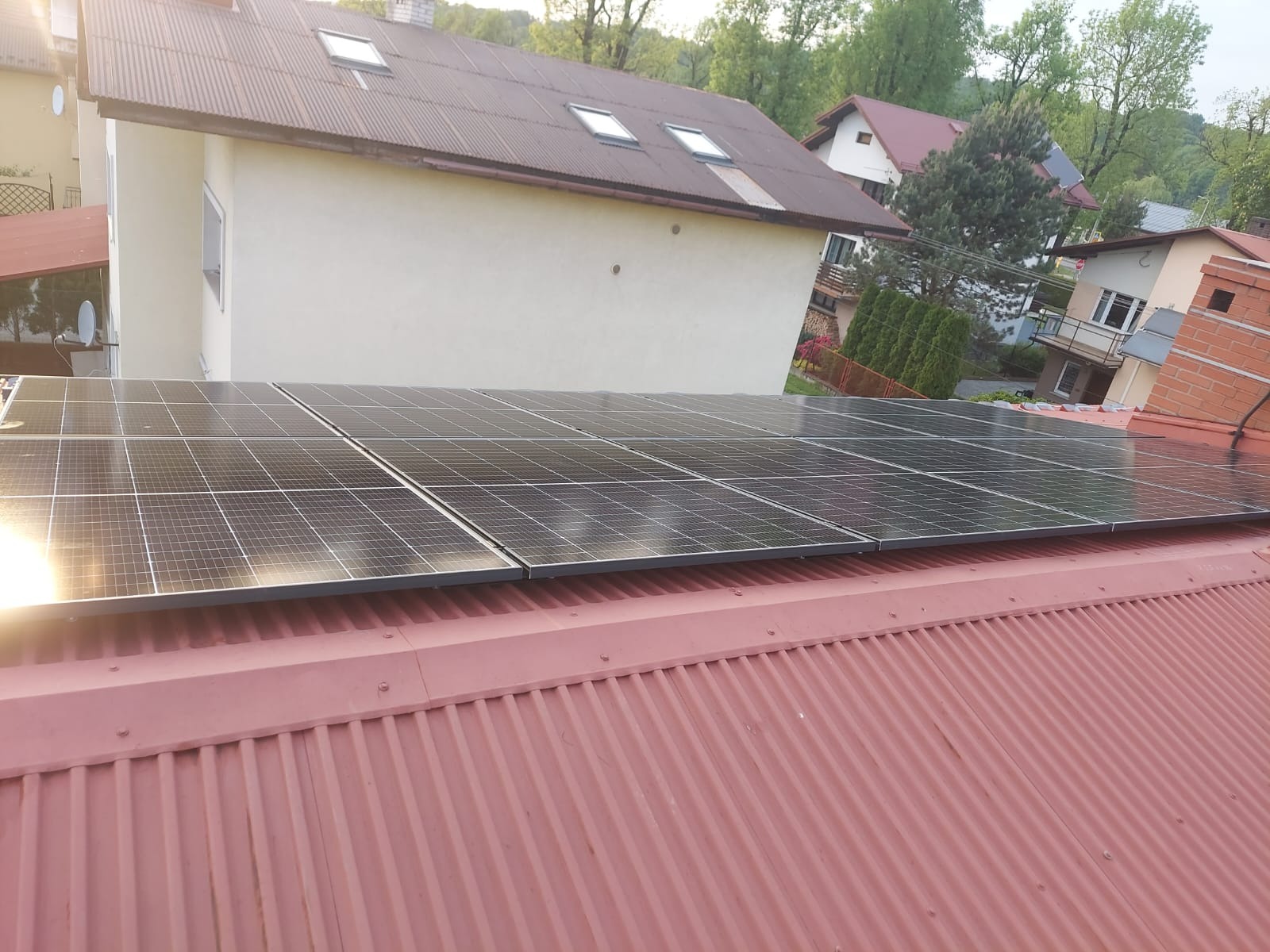 Panele słoneczne na czerwonym, blaszanym dachu domu jednorodzinnego w słoneczny dzień. Widok z góry, z bliska, z widocznymi detalami konstrukcji dachu i paneli.