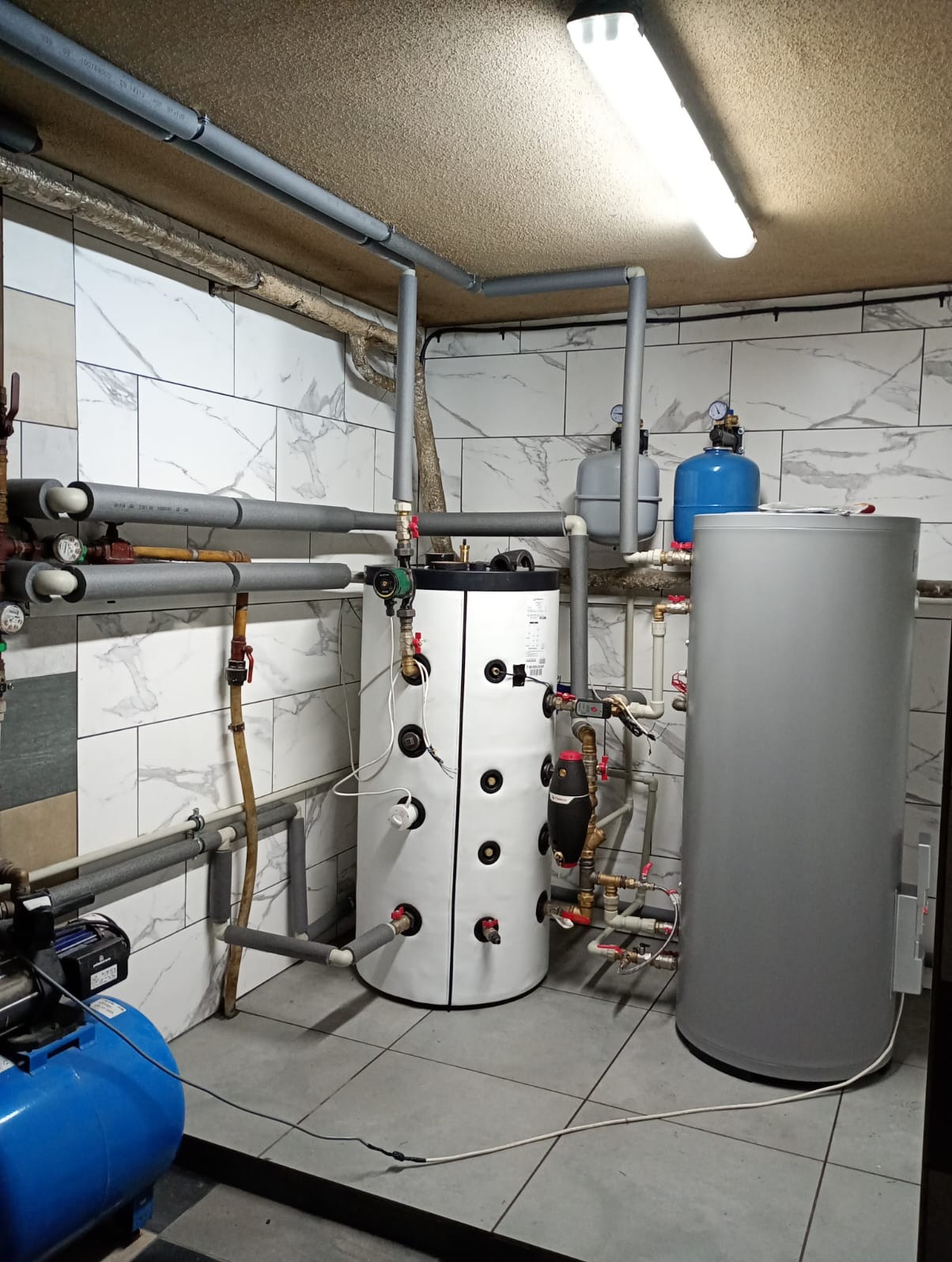 Kotłownia z dwoma zbiornikami, rurami w izolacji i białymi płytkami imitującymi marmur na ścianach. Widoczne elementy instalacji grzewczej.