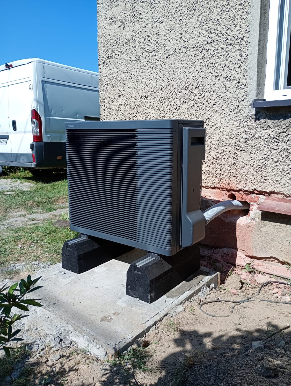 Szara pompa ciepła Panasonic Aquarea zamontowana na betonowym fundamencie przy ścianie budynku z szorstką elewacją. Widoczny biały bus w tle.