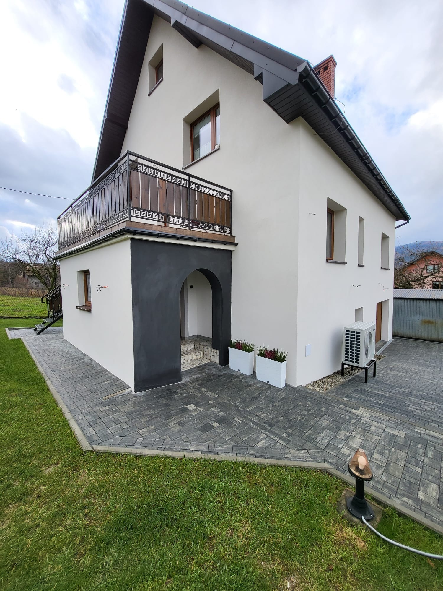 Dwukondygnacyjny dom z balkonem, wykończony w bieli i szarości, z widocznym klimatyzatorem i brukowanym podjazdem. Uwagę zwraca ciemnoszara wnęka wejściowa.