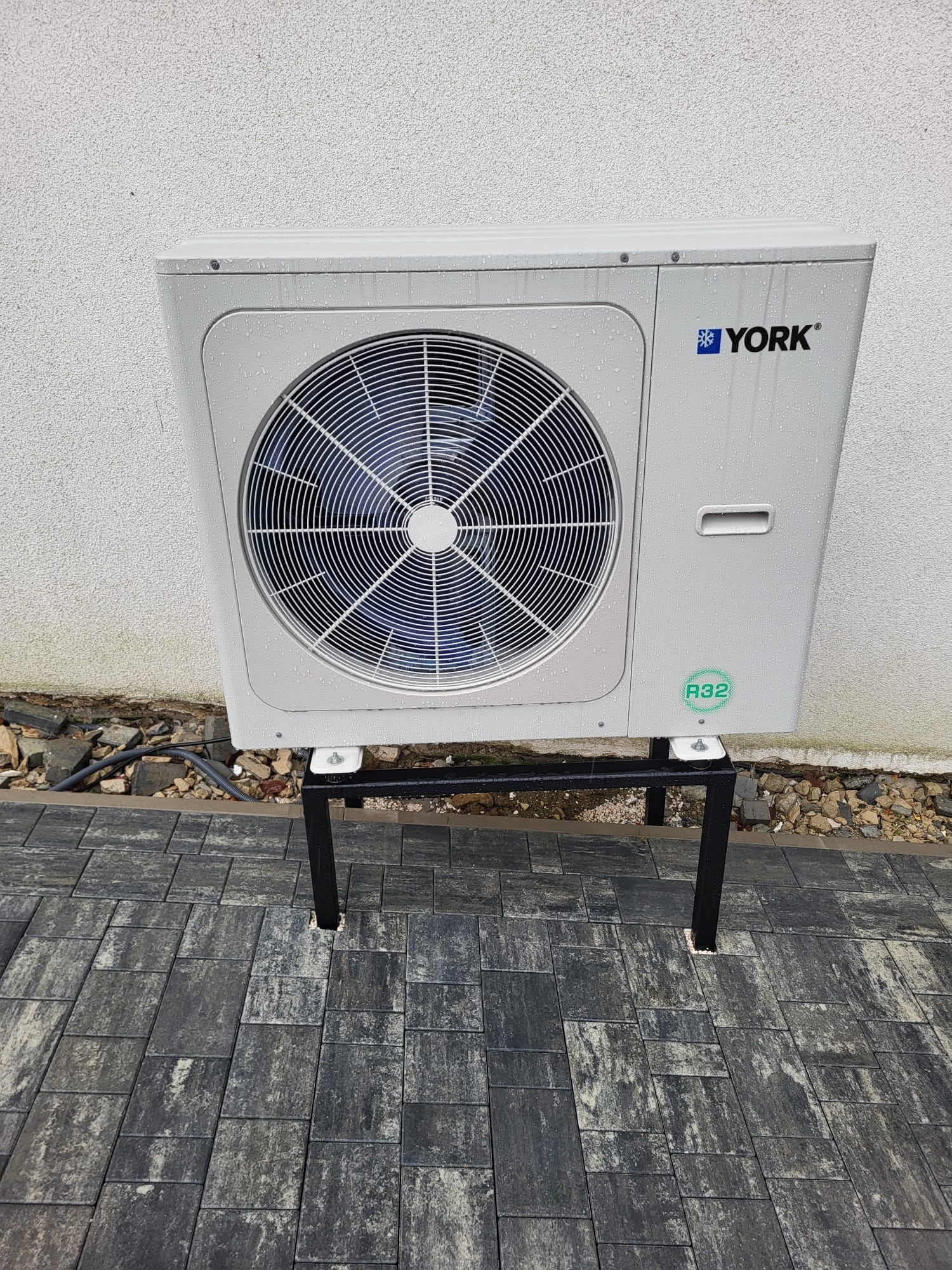 Biała jednostka zewnętrzna klimatyzacji York na czarnym stelażu, ustawiona na szarej kostce brukowej przy białej ścianie. Widoczne krople wody na obudowie.