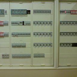 KAMKOP Pałuki Kamil Kopicki - Rozbudowana, trójsegmentowa tablica elektryczna z licznymi bezpiecznikami, wyłącznikami i modułami, umieszczona w białej obudowie w jasnym pomieszczeniu.