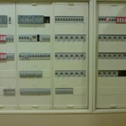 Rozbudowana, trójsegmentowa tablica elektryczna z licznymi bezpiecznikami, wyłącznikami i modułami, umieszczona w białej obudowie w jasnym pomieszczeniu.