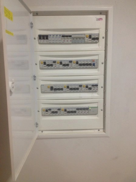 Otwarta skrzynka elektryczna z czterema rzędami bezpieczników i przełączników, zamontowana na białej ścianie.