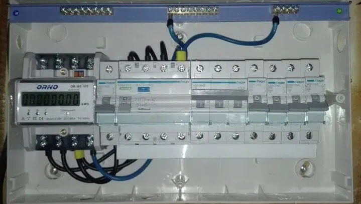 Wewnętrzny widok rozdzielnicy elektrycznej z zamontowanym licznikiem energii i zabezpieczeniami, widoczne przewody i listwy zaciskowe.