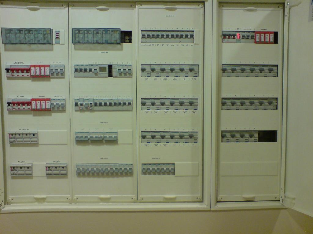 Rozbudowana, trójsegmentowa tablica elektryczna z licznymi bezpiecznikami, wyłącznikami i modułami, umieszczona w białej obudowie w jasnym pomieszczeniu.