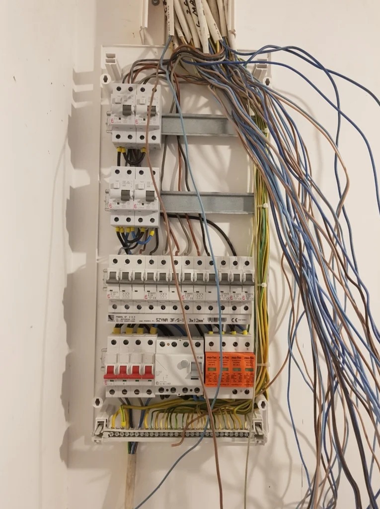 Rozdzielnica elektryczna z widocznymi bezpiecznikami, przewodami i listwami zaciskowymi na białej ścianie.