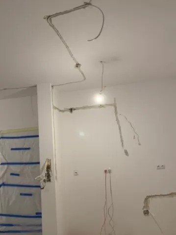 Wnętrze w trakcie remontu z widocznymi przewodami elektrycznymi poprowadzonymi po suficie i ścianach, zabezpieczona folią strefa malarska.