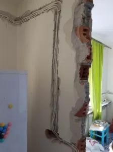 Kable elektryczne w białej osłonie prowadzone w bruzdach wyciętych w ścianie podczas remontu, widoczne fragmenty cegieł, w tle lodówka i zasłona okienna.