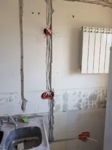 Ściana w trakcie remontu z wyżłobieniami na przewody elektryczne, widoczne puszki instalacyjne z wystającymi kablami, fragment zlewu i grzejnika.