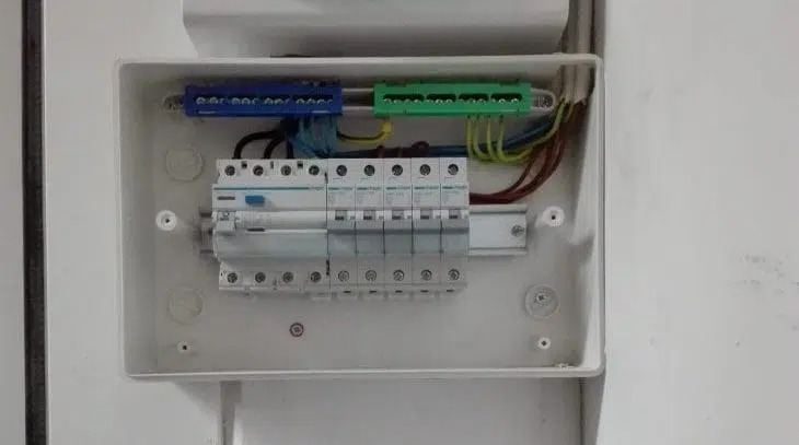 Otwarta skrzynka elektryczna z zainstalowanymi bezpiecznikami, widoczne przewody w kolorowych listwach zaciskowych, z bliska, zamontowana na białej ścianie.