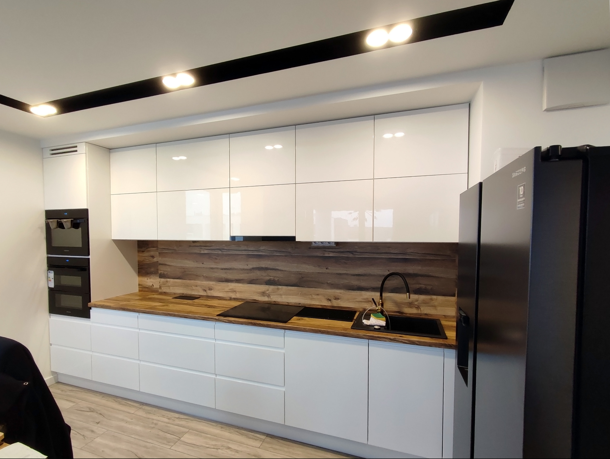 Nowoczesna kuchnia: białe, lakierowane szafki, drewniany blat i panel ścienny, czarna armatura i lodówka Samsung.