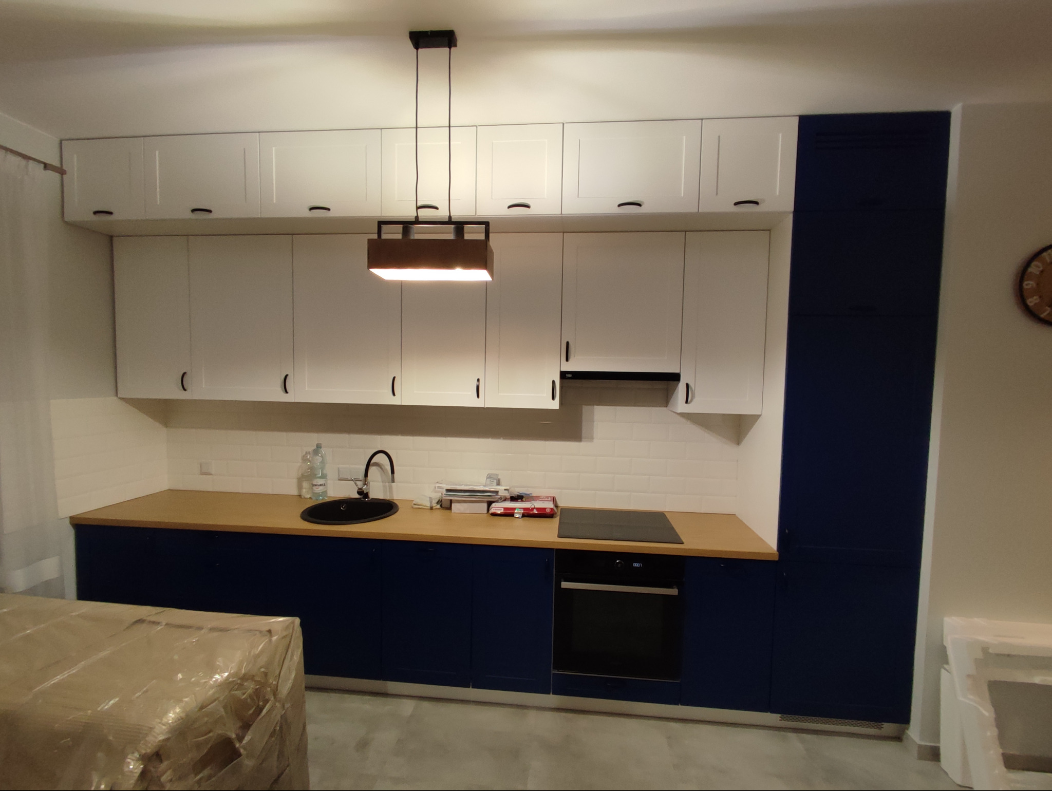 Nowoczesna kuchnia z białymi szafkami górnymi i granatowymi dolnymi, drewniany blat, wisząca lampa, białe płytki na ścianie, częściowo zasłonięty karton na pierwszym planie.