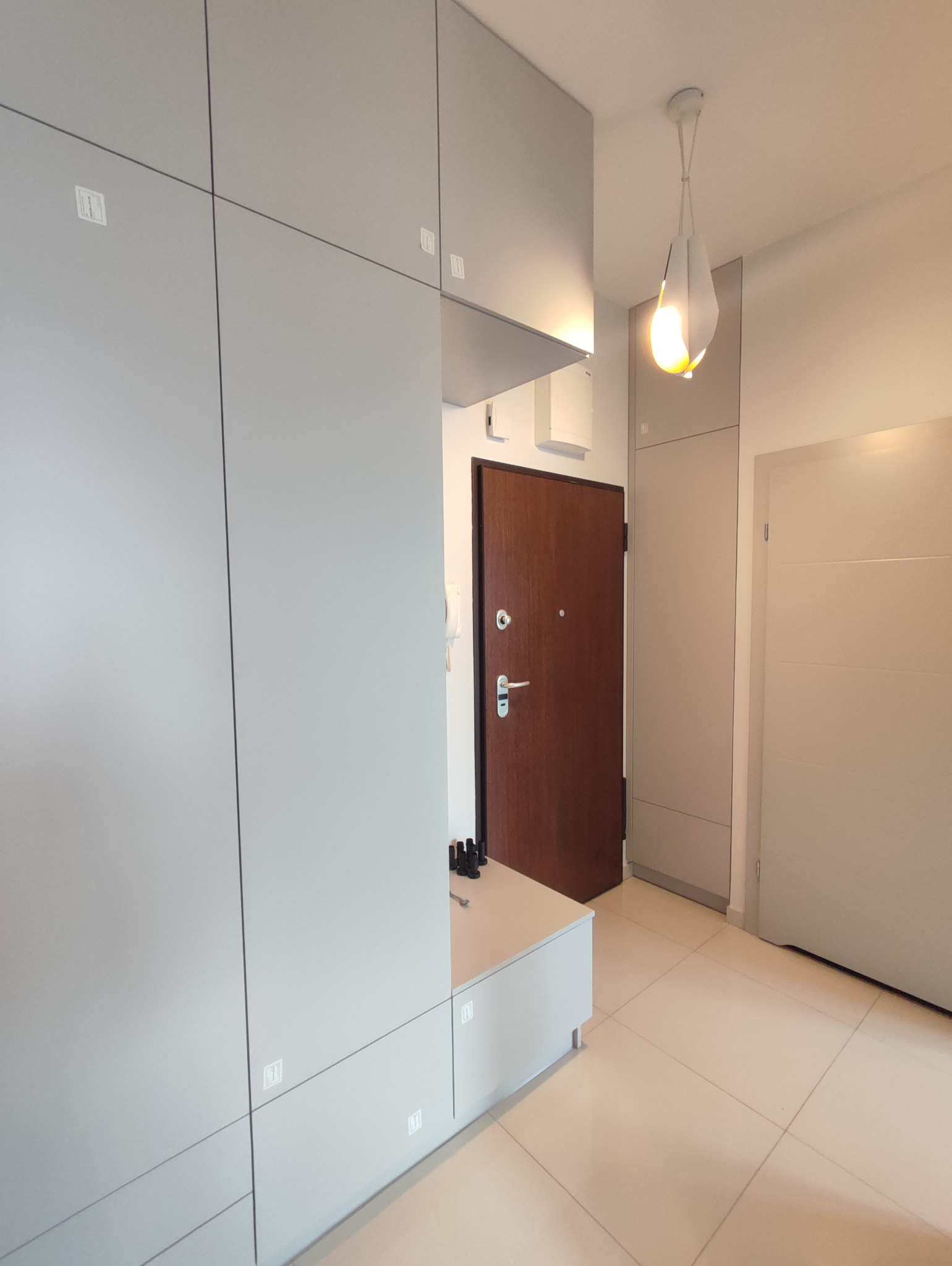 Szara zabudowa meblowa w przedpokoju z drewnianymi drzwiami wejściowymi i nowoczesną lampą wiszącą.