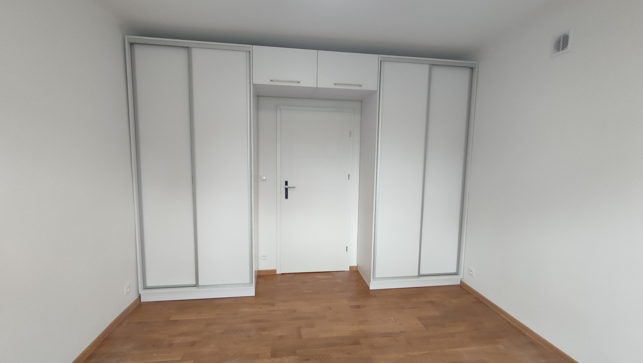 Biała zabudowa meblowa z szafami przesuwnymi i szafkami nad drzwiami w minimalistycznym wnętrzu z parkietem.