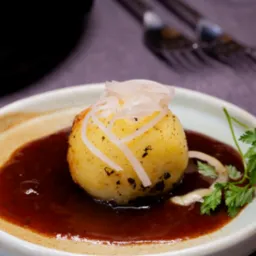 Ziemniaczana kula z brązowym sosem, posypana białymi paseczkami, udekorowana zieloną gałązką pietruszki, na ceramicznym talerzu.