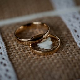 Pro SmArt Studio Adam Kowal - Dwa złote obrączki ślubne spoczywają na jutowym podłożu z białą koronkową wstążką, pomiędzy nimi mała biała ozdoba w kształcie serca.